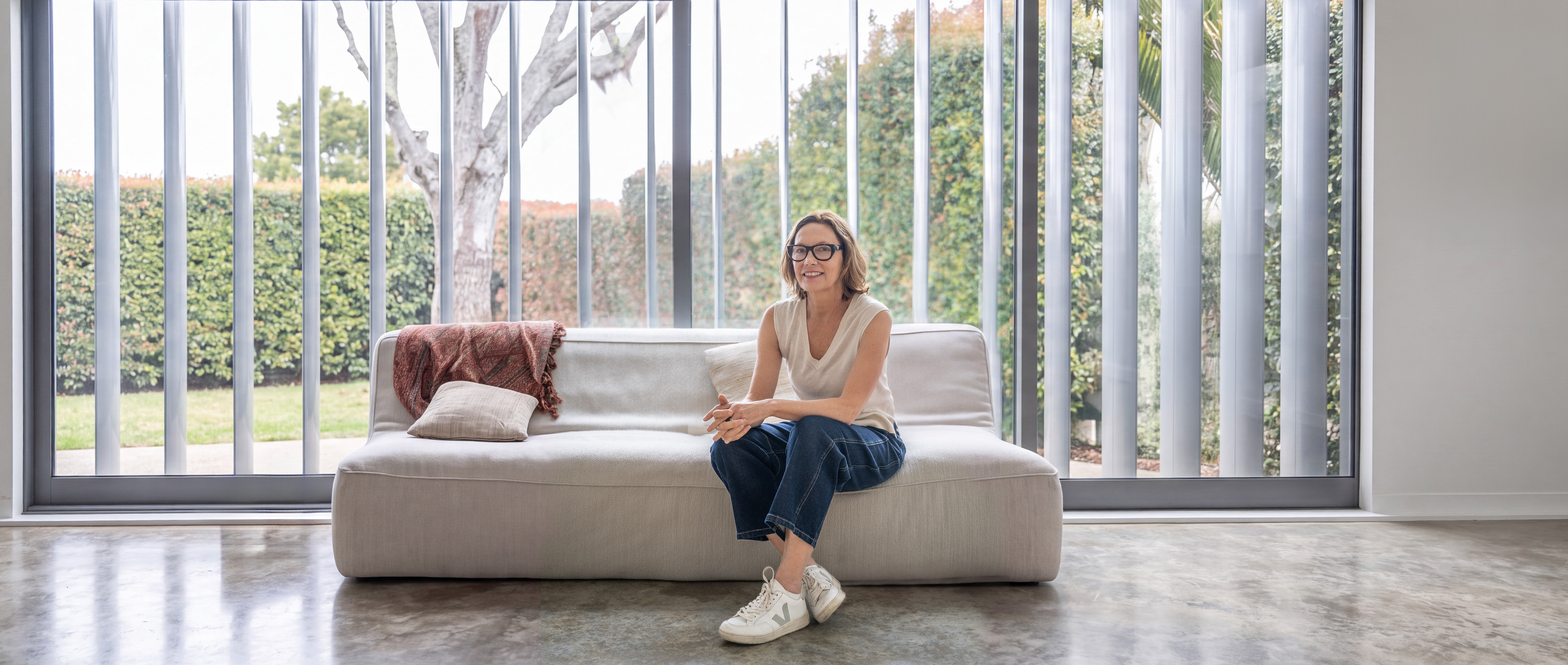 Tina Bull seated on a sofa in a bright studio, with tall windows and a garden behind her