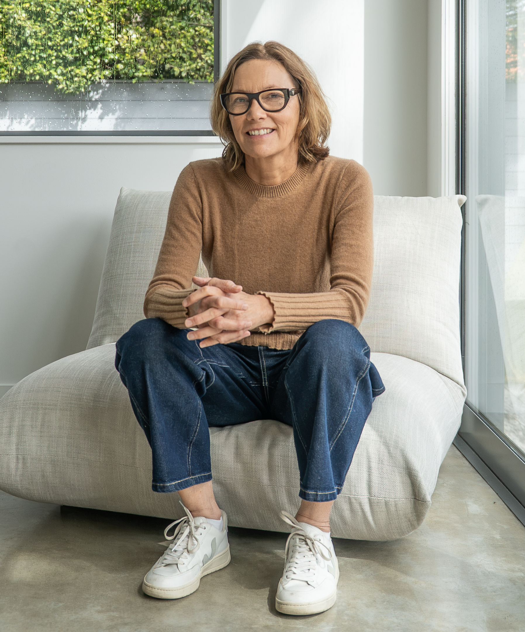 Tina Bull, smiling, seated by a window in a camel sweater and glasses